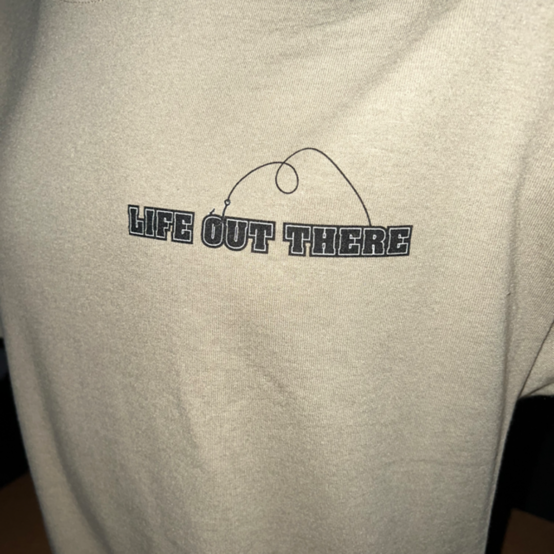 A close-up of the Unisex The Hooker T Shirt in Khaki/Black by Life Out There Apparel, showcasing the phrase "LIFE OUT THERE" boldly printed on it. Above the text is a simple line drawing that encapsulates fishing, featuring a rod and line elegantly arched over the words.