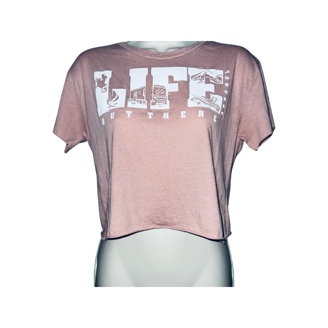 The OG Women's Crop T Shirt - Desert Pink/White by Life Out There Apparel is presented on a mannequin, featuring the word LIFE in white uppercase letters with each letter illustrating a unique camping scene. The phrase OUT THERE is printed below in a smaller font.