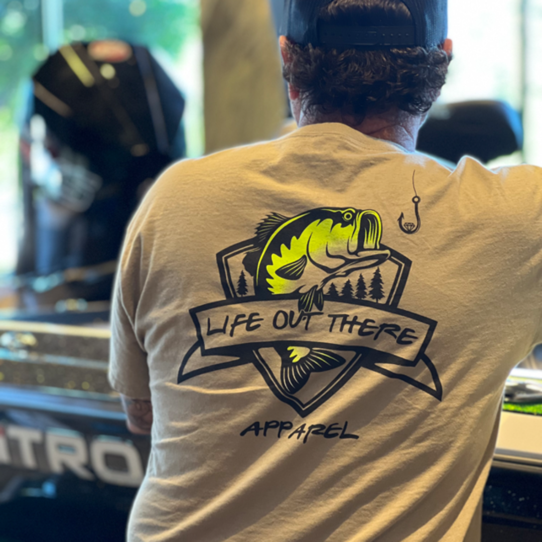 A man wearing the Unisex The Hooker T Shirt in Khaki/Black by Life Out There Apparel stands near a boat. He's in a store or showroom with large windows in the background, showcasing the perfect garment for any fishing enthusiast.