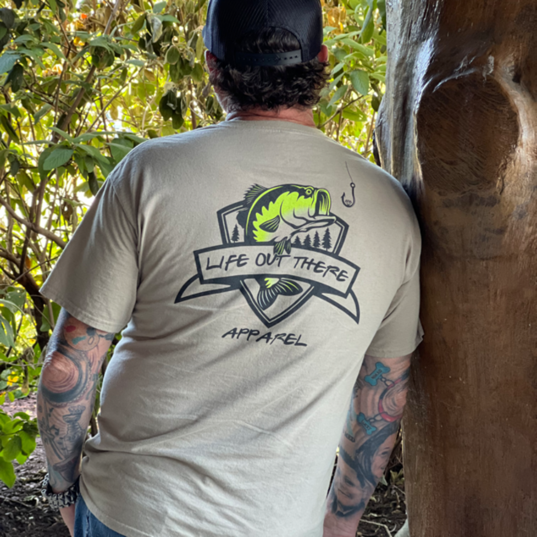 A person wearing a khaki and black Unisex The Hooker T-Shirt from Life Out There Apparel, featuring a graphic of a fish and fishing hook, stands against a tree. This stylish garment complements their tattooed arms and black cap, while the lush greenery in the background adds a touch of nature-inspired elegance.