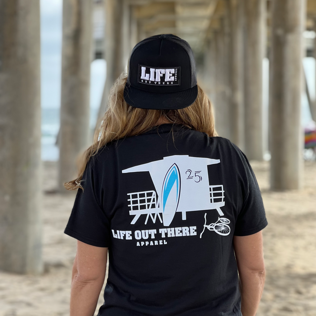 A person dressed in a black Unisex The JJ T Shirt by Life Out There Apparel and a matching cap stands under a pier, facing away. The T-shirt sports a surfboard design with the number 25, while the cap spells out LIFE. The sandy beach suggests an idyllic summer vacation setting, with the pier's structure prominently visible in the background.