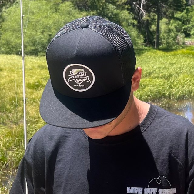 A person is wearing The Hooker Snapback Trucker Hat in black by Life Out There Apparel, featuring a logo and a bass fish, paired with a matching black T-shirt. They stand outdoors against a green grassy backdrop, holding a fishing pole.