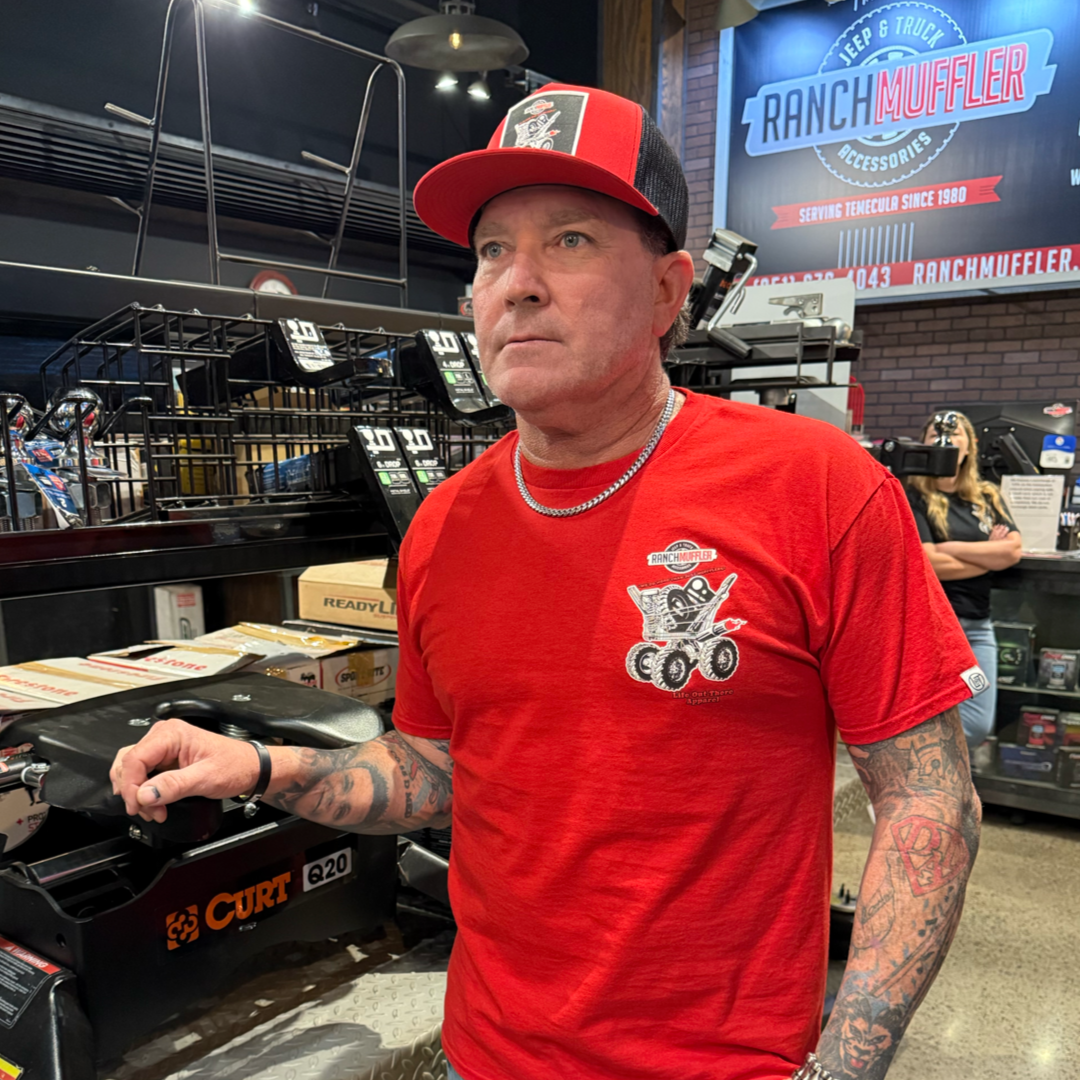 A tattooed man wears the Life Out There Apparel Unisex "We Do More Than Just Mufflers" Tee in red and a matching cap, standing in an automotive shop with Jeep & Truck Accessories, mufflers, and lift kits by a RANCH MUFFLER sign.