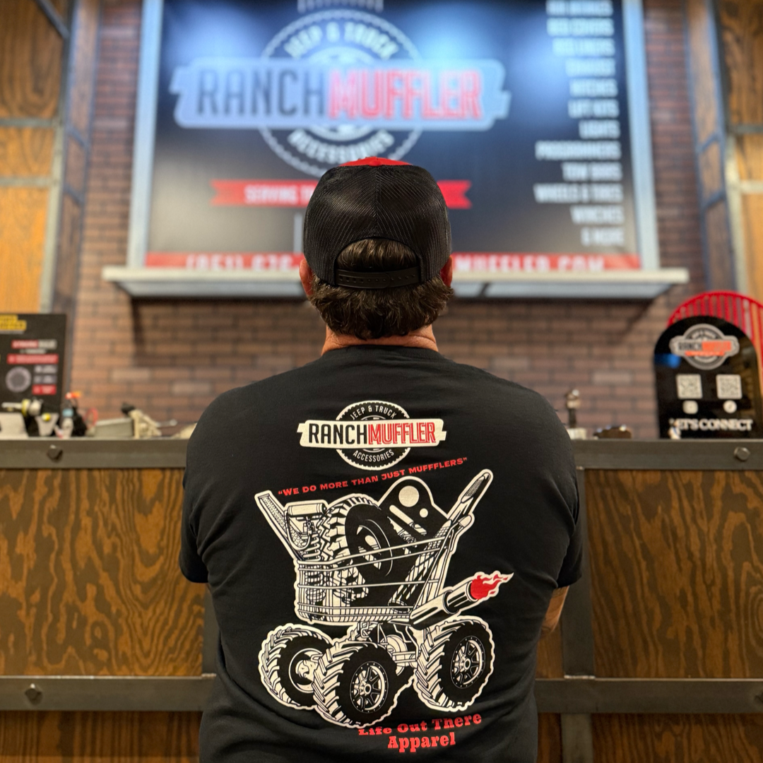 A man wearing a black "We Do More Than Just Mufflers" tee by Life Out There Apparel stands with his back to the camera, facing a shop counter and a large Ranch Muffler sign—your go-to for mufflers and quality truck accessories.