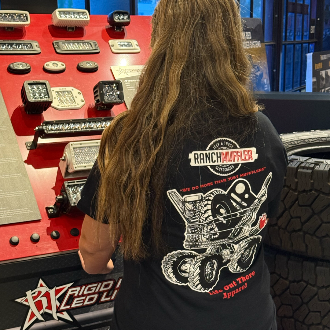 A woman with long brown hair wears a black "We Do More Than Just Mufflers" tee by Life Out There Apparel, standing in front of off-road LED lights and Jeep accessories. The shirt features a vehicle and tire-themed graphic.