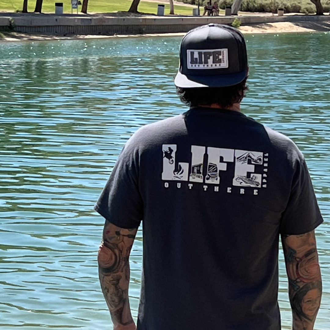 A person with tattooed arms stands by a body of water, wearing a black cap and the Unisex The OG T Shirt in Gray/White from Life Out There Apparel. Nearby, dirt bikes are propped against trees, adding to the scene filled with grass, benches, and the promise of adventure.