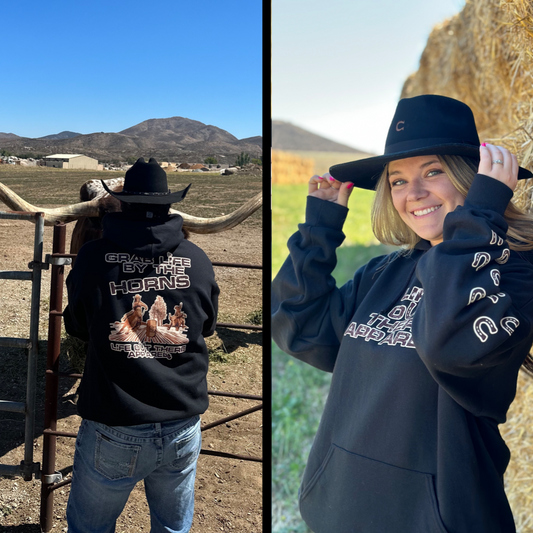 Life Out There Apparel “Grab Life By The Horns” Black Unisex Hoodie has a girl barrel racer getting ready to turn around a barrel with a guy riding a bull behind her.  This shows a guy wearing the black hoodie, standing in front of a bull & a girl wearing the black hoodie with a cowboy hat on.   