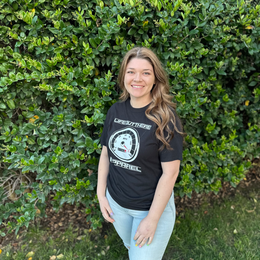 Life Out There Apparel “On My Way” Black Unisex tee shirt is the 1 year anniversary design that has a big speedometer on it - pegged at 120 mph & the odometer is rolling over 1 Year. This photo shows a woman showing the design on the front of the of the shirt standing in front of a wall of green bushes. 