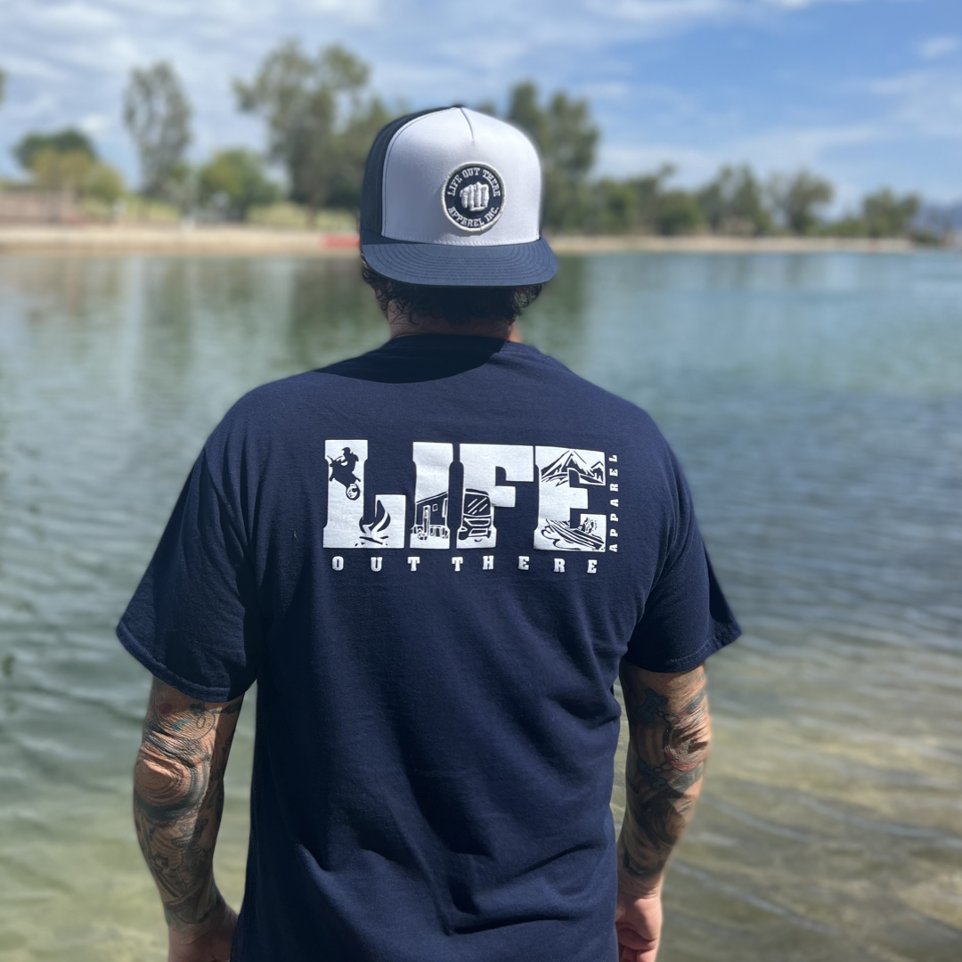 A tattooed individual stands beside a tranquil lake beneath a clear blue sky, donned in a navy Unisex The OG T Shirt by Life Out There Apparel and a baseball cap. They appear prepared for camping escapades with trees and vast landscape extending into the background.