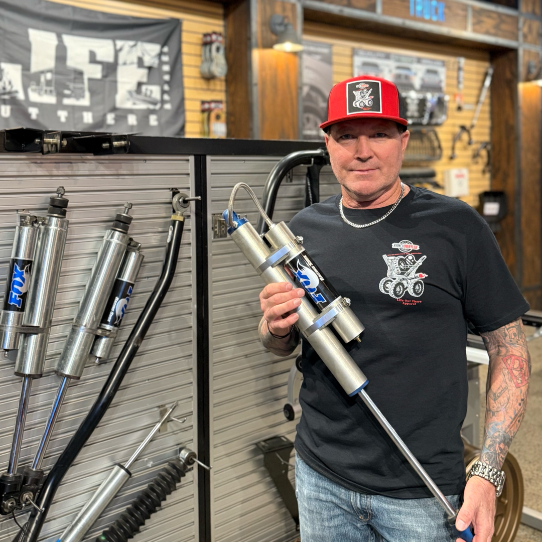 A man in the Life Out There Apparel Unisex "We Do More Than Just Mufflers" Black Tee stands indoors holding a large shock absorber. Behind him, Jeep and truck accessories, shock absorbers, mufflers, and motorsport gear line the wall and workbench.