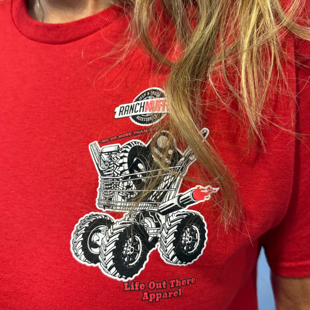 Wearing the red "Unisex - We Do More Than Just Mufflers Tee" by Life Out There Apparel, featuring a Ranch Muffler logo and tires in a cart on monster truck wheels—perfect for muffler and Jeep & Truck Accessories fans.