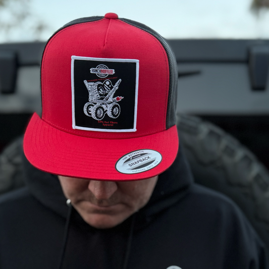 A person wearing the Life Out There Apparel "We Do More Than Just Mufflers" Flat Bill SnapBack Trucker Hat in red and black looks down, with a blurred vehicle tire hinting at Jeep accessories or lift kits in the background.