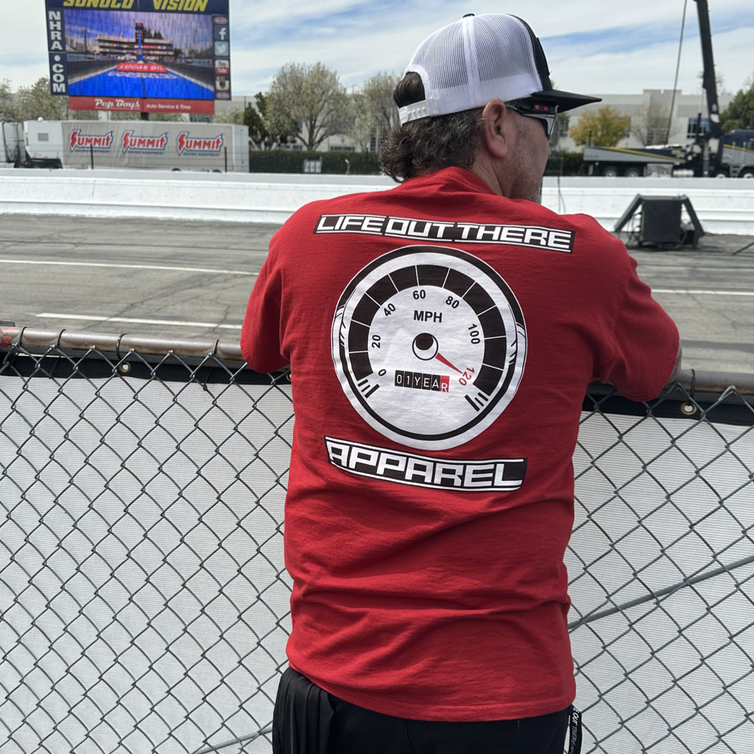 The Crimson Red Life Out There Apparel “On My Way” Unisex tee shirt is the 1 year anniversary design that has a big speedometer on it - pegged at 120 mph & the odometer is rolling over 1 Year. This is a photo of a man standing at a fence at the edge of a drag strip showing the back of the shirt. 