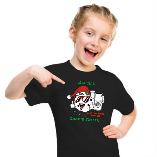A smiling girl in the Life Out There Apparel Unisex Toddler Official Cookie Tester T Shirt - Black, featuring a festive cartoon cookie, sunglasses, Santa hat, and 2024 Official Cookie Tester—perfect for delivering Christmas cookies to Santa.