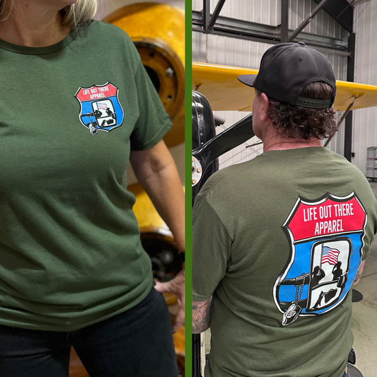Military Green Unisex tee shirt from Life Out There Apparel “Leave It Behind” The design has a truck side mirror with the reflection of a male, female & K9 soldiers saluting the American flag (inside of an interstate sign) - bringing awareness to P. T. S. D. (post traumatic stress disorder) This photo shows a women showing the front of the shirt & a man showing the back design standing next to an old war airplane. 