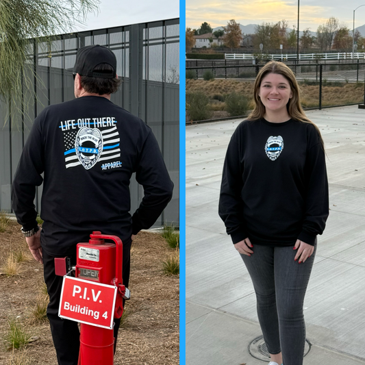 Two people wear Life Out There Apparels black Unisex Back The Blue long sleeve tee with a blue-striped flag design. The man, near a red P.I.V Building 4 sign, turns away while the woman smiles at the camera, posing on a paved surface outdoors.