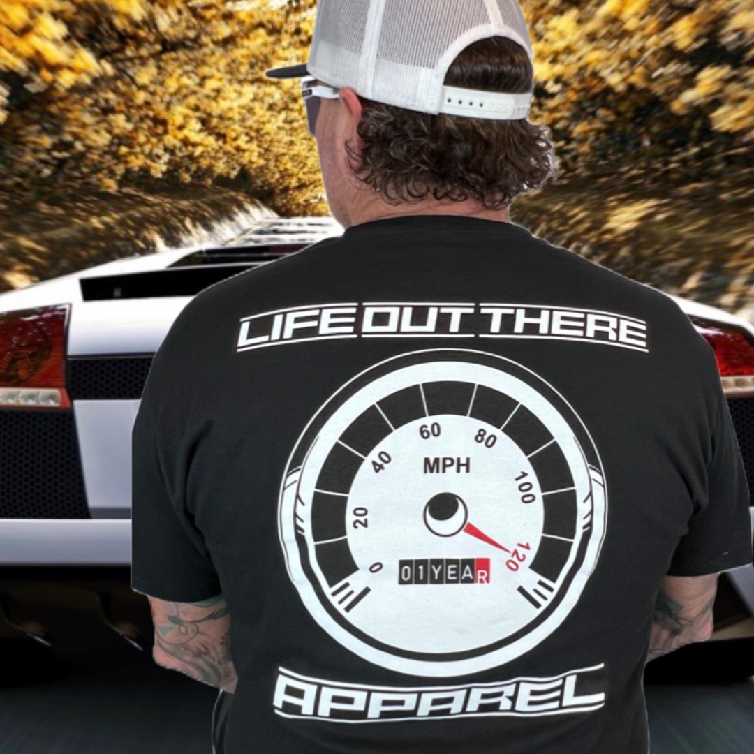 Life Out There Apparel “On My Way” Black Unisex tee shirt is the 1 year anniversary design that has a big speedometer on it - pegged at 120 mph & the odometer is rolling over 1 Year. This photo shows a man showing the design on the back of the shirt standing behind a white sports car.   