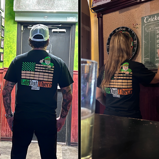 Life Out There Apparel “Capt. Patrick” Unisex tee shirt in black is a collaboration of an American flag with an Irish twist and a leprechaun boat Captain looking on. This is a photo,of a man & women wearing the shirt in a pub. 