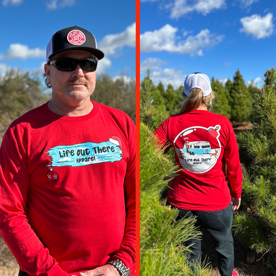 Life Out There Apparel “December 26th” red Unisex long sleeve tee shows Santa Clause camping in the desert, in a travel trailer, while relaxing after the big day and burning his Christmas tree, all inside a Christmas ornament. A women showing the back of the shirt & a man showing the front of the shirt. 