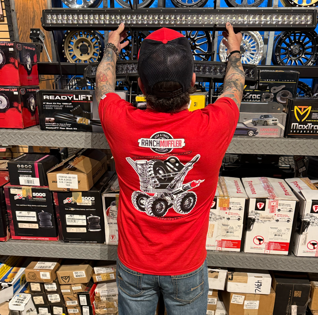 Someone wearing a red "Unisex - We Do More Than Just Mufflers Tee" by Life Out There Apparel is reaching for a shelf in an auto parts store, with boxes of mufflers and Jeep & Truck Accessories around them, back to the camera.