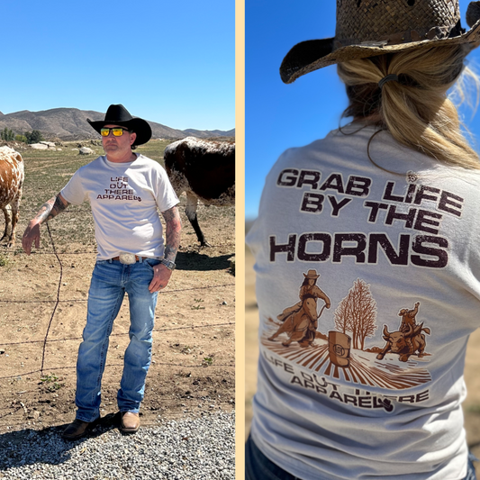 Life Out There Apparel Sandstone colored “Grab Life By The Horns” Unisex tee shirt has a girl barrel racer getting ready to turn around a barrel with a guy riding a bull behind her.  This shows a guy showing the front of the shirt standing in front of some long horn bulls & a women showing the back of the tee. 