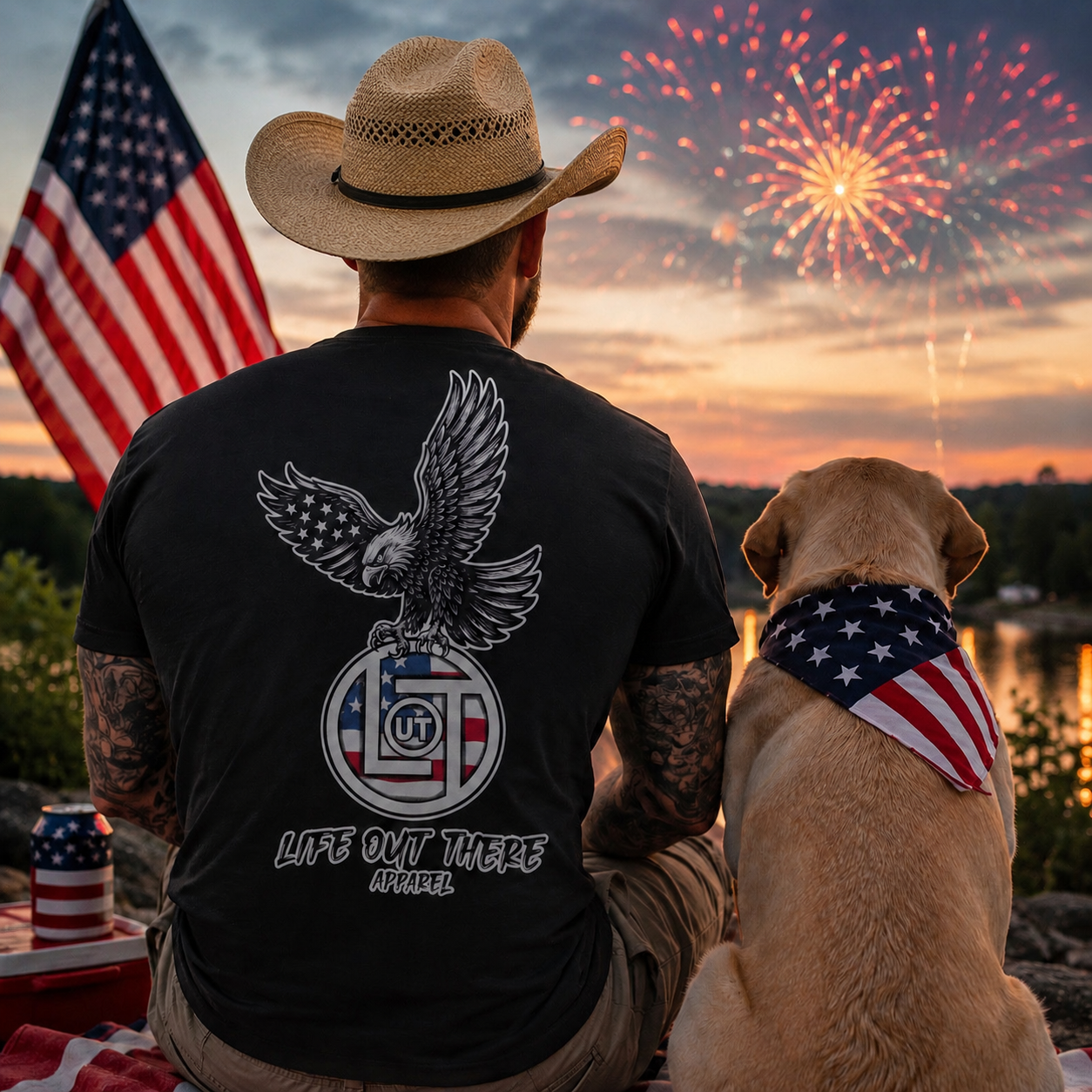 A man in a cowboy hat and his dog, both wearing American flag accessories, sit outdoors at sunset watching fireworks. He sports Life Out There Apparel’s Flight of Freedom Tee - Black, featuring an eagle and patriotic design as a flag waves nearby.