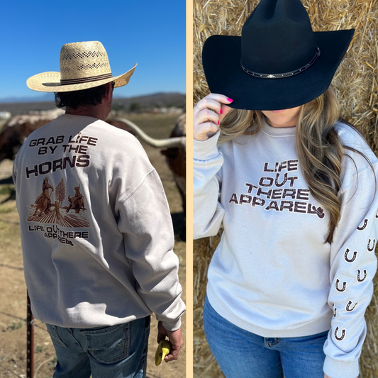 Life Out There Apparel Sandstone colored “Grab Life By The Horns” Unisex Crew Neck Sweatshirt has a girl barrel racer getting ready to turn around a barrel with a guy riding a bull behind her.  This shows a guy wearing the sweatshirt standing in front of a bull & a girl wearing the sweatshirt with a cowboy hat on.   