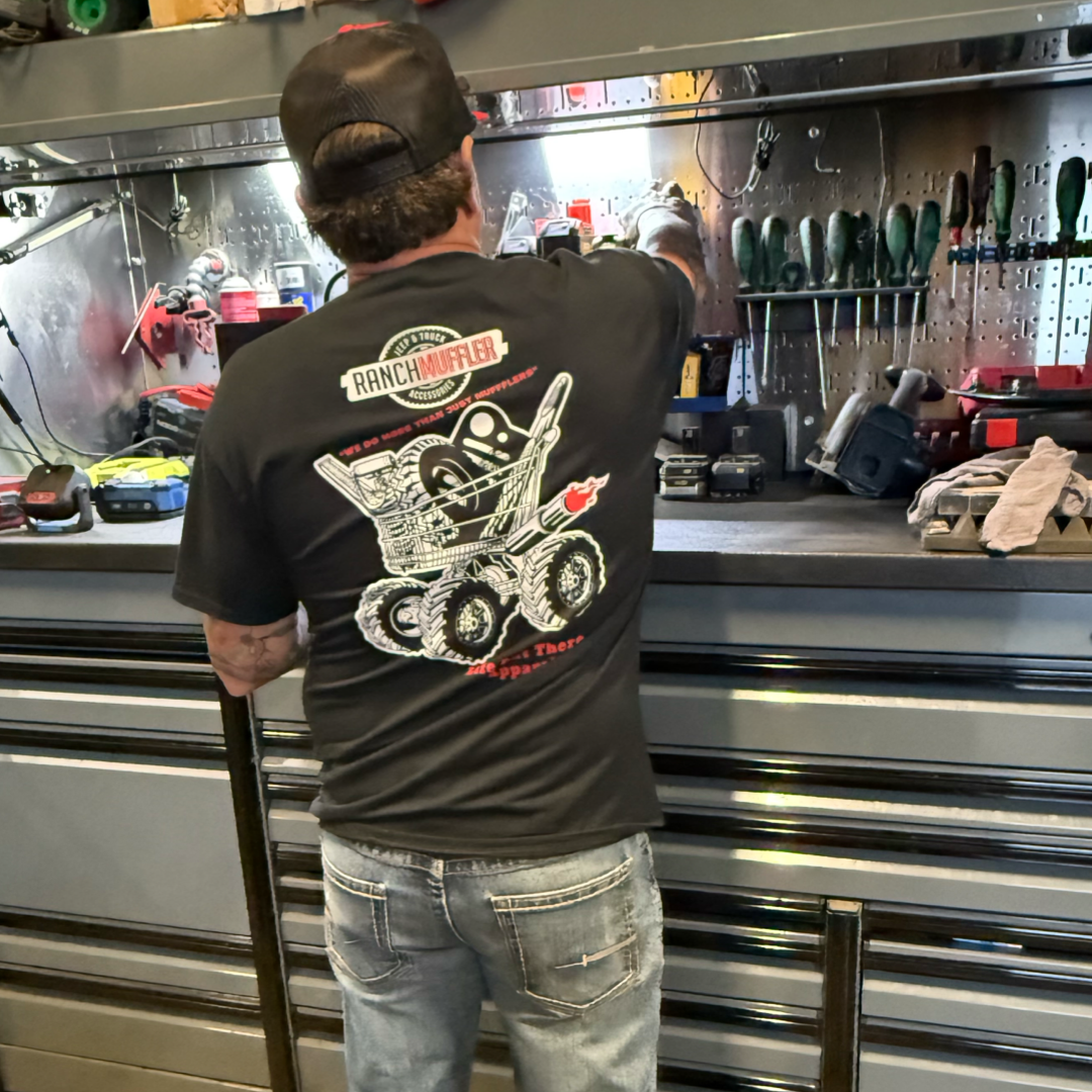 A man in a black "Unisex - We Do More Than Just Mufflers Tee" by Life Out There Apparel works at a tool bench, reaching for tools on a metallic pegboard, with truck accessories and equipment organized nearby.
