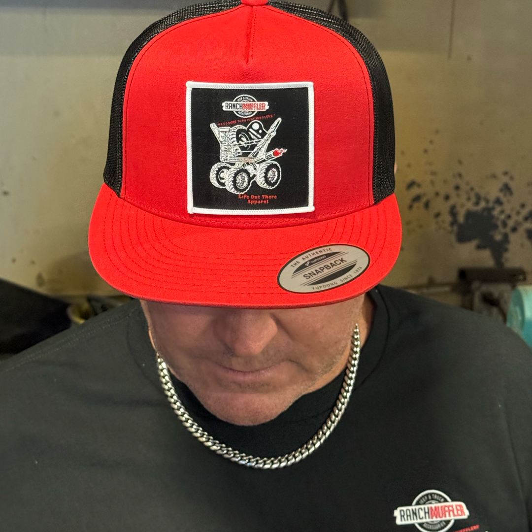 A man wearing a Life Out There Apparel "We Do More Than Just Mufflers" Red/Black Flat Bill SnapBack Trucker Hat with an embroidered eagle, text, and red heart looks down, pairing it with a black shirt and silver chain.