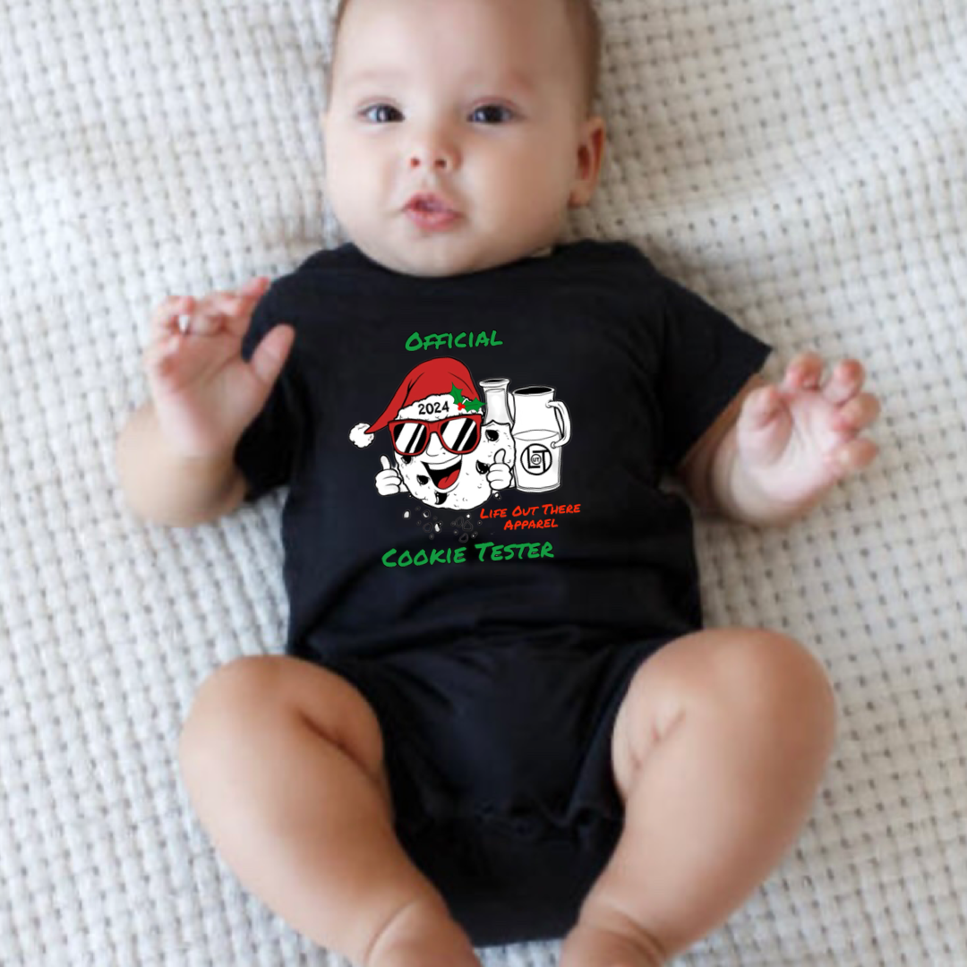 Black short sleeve baby shirt with a cartoon cookie wearing a Santa hat and sunglasses labeled "2024," alongside glass milk cartons. Text reads, "Life Out The Apparel / official cookie tester”. 