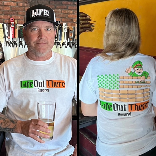 Life Out There Apparel “Capt. Patrick” Unisex tee shirt in white is a collaboration of an American flag with an Irish twist and a leprechaun boat Captain looking on. This is a photo of a man with a beer & a women wearing the shirt in a pub. 