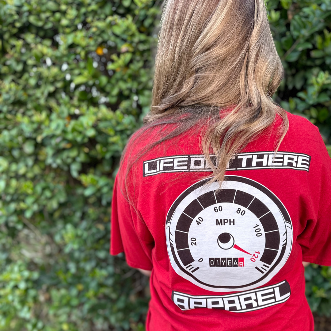 The Crimson Red Life Out There Apparel “On My Way” Unisex tee shirt is the 1 year anniversary design that has a big speedometer on it - pegged at 120 mph & the odometer is rolling over 1 Year. This is a photo of a young lady showing the back of the shirt standing in front of a row of green bushes. 