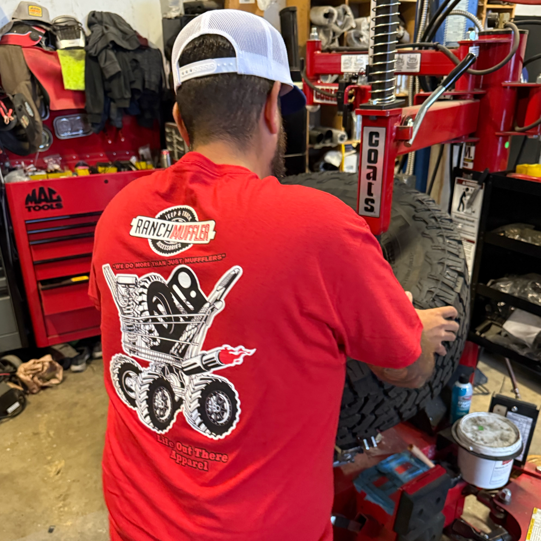 Wearing the Life Out There Apparel Unisex "We Do More Than Just Mufflers" Tee in red, a man equips a large off-road tire with a mounting machine in a cluttered garage filled with Jeep & Truck Accessories, mufflers, and lift kits.