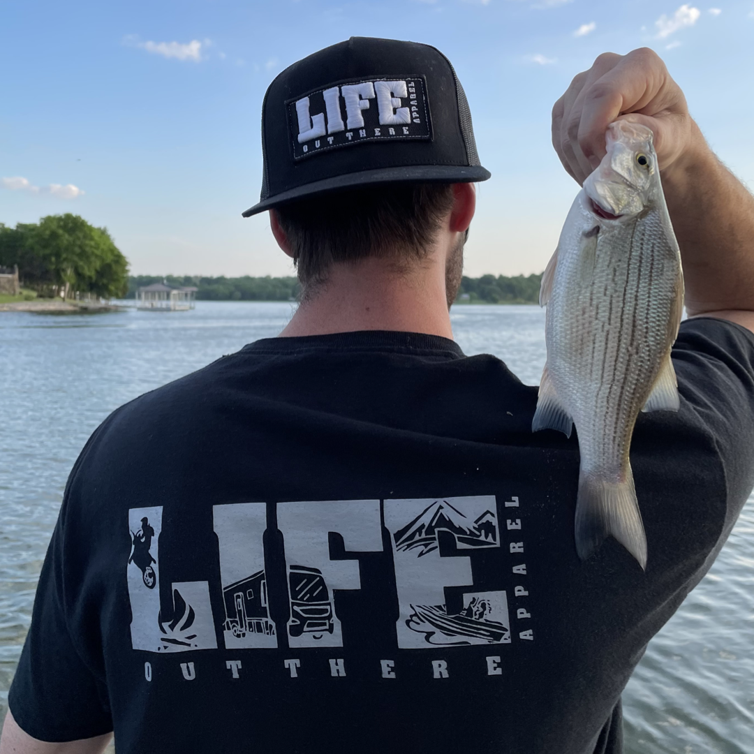 Wearing a black and grey Unisex The OG T Shirt by Life Out There Apparel, a person holds a fish by the serene lakeside, surrounded by trees and buildings under a partly cloudy sky, encapsulating the spirit of an adventurous day with nearby snowboarding.