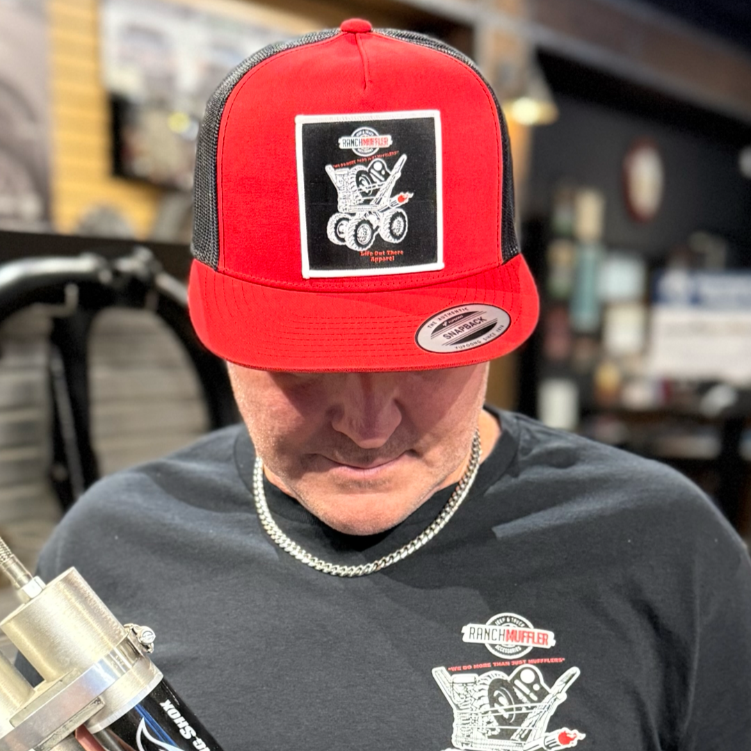 A person wearing the Life Out There Apparel "We Do More Than Just Mufflers" Flat Bill SnapBack Trucker Hat in Red/Black and a matching tee with Ranch Muffler logos looks down, holding a metallic muffler indoors.