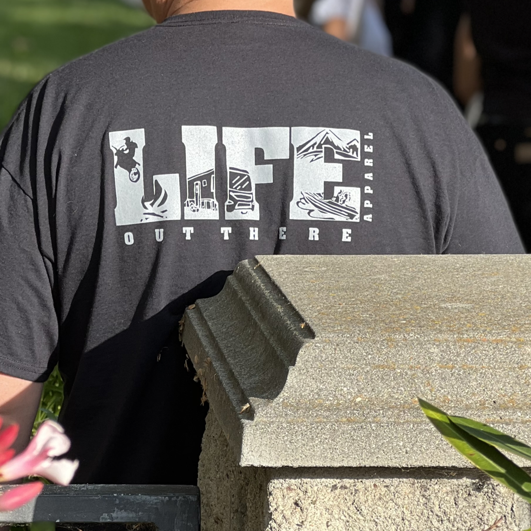 A person wearing the Unisex The OG T Shirt in Black/Grey by Life Out There Apparel stands outdoors near a stone structure. The t-shirt is adorned with small illustrations, including a surfer and mountains, capturing the essence of camping adventures. Greenery and pink flowers enhance the charm of the scene.