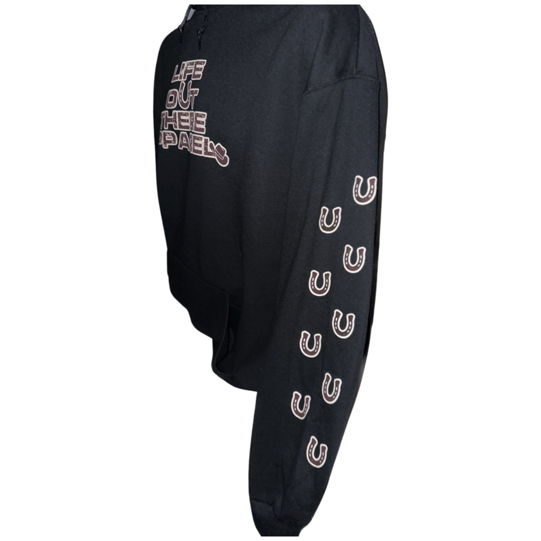 Life Out There Apparel “Grab Life By The Horns” Black Unisex Hoodie has a girl barrel racer getting ready to turn around a barrel with a guy riding a bull behind her.  This shows the sleeve of the black hoodie that has horseshoes up the sleeve.  
