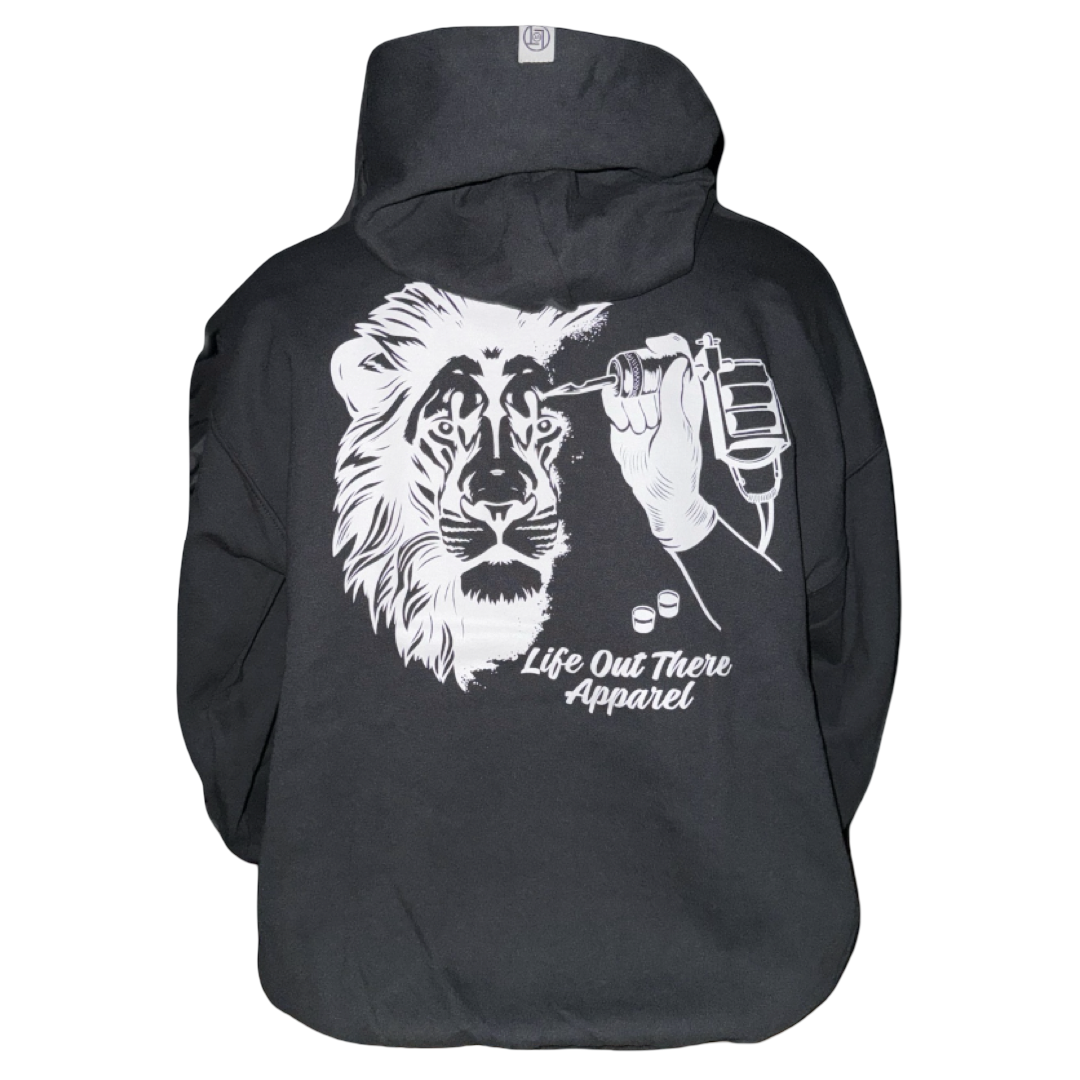 The Unisex - Not All There Hoodie in black by Life Out There Apparel features a soft cotton build, tagless comfort, and a striking graphic of a hand tattooing a lion’s face with “Life Out There Apparel” text below the artwork.