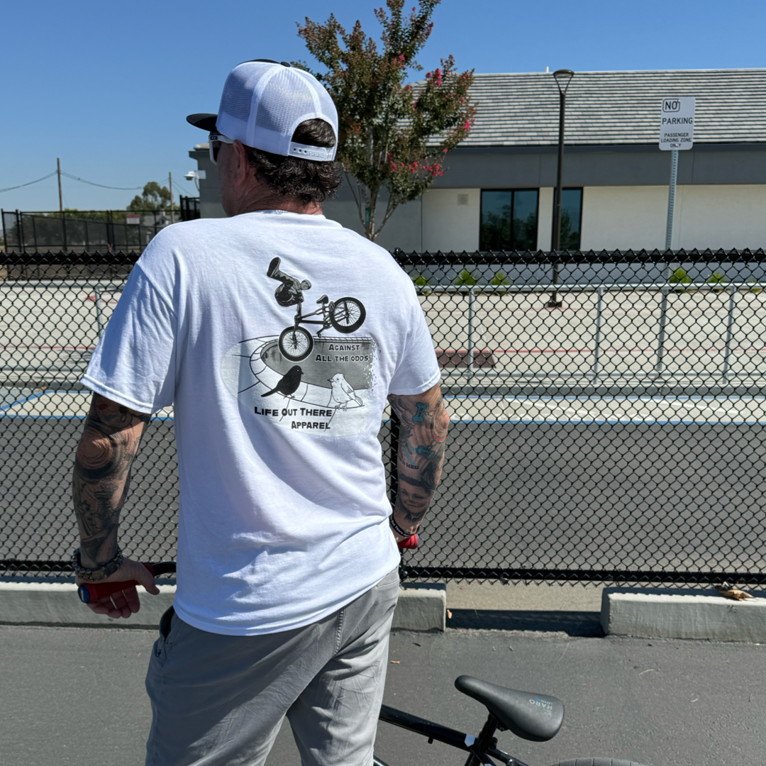 A man in a white Unisex - Against All The Odds / The Ride Tee by Life Out There Apparel and a cap stands next to a BMX bike on a sunny street, his shirt’s bold graphic inspired by BMX legend John Buultjens.