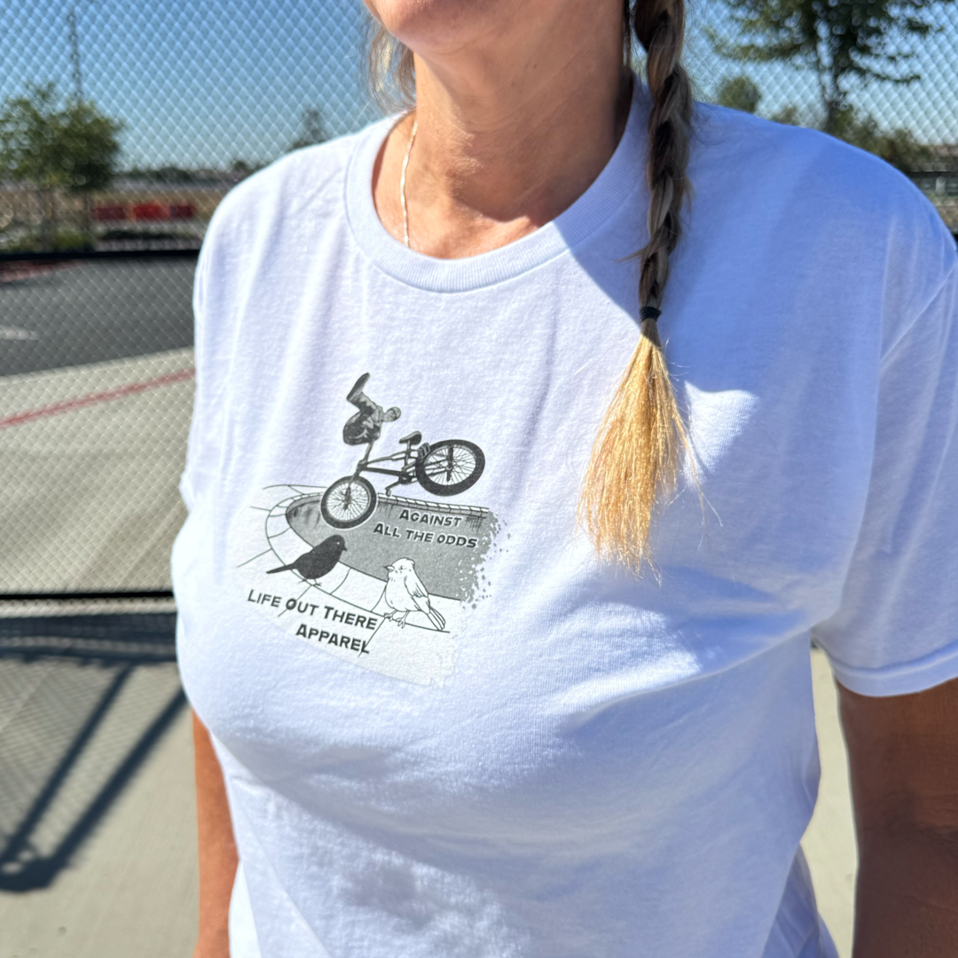 A woman with a blonde braid wears the white Unisex Against All The Odds / The Ride Tee by Life Out There Apparel, featuring BMX legend John Buultjens, while standing outdoors near a fence on a sunny day.