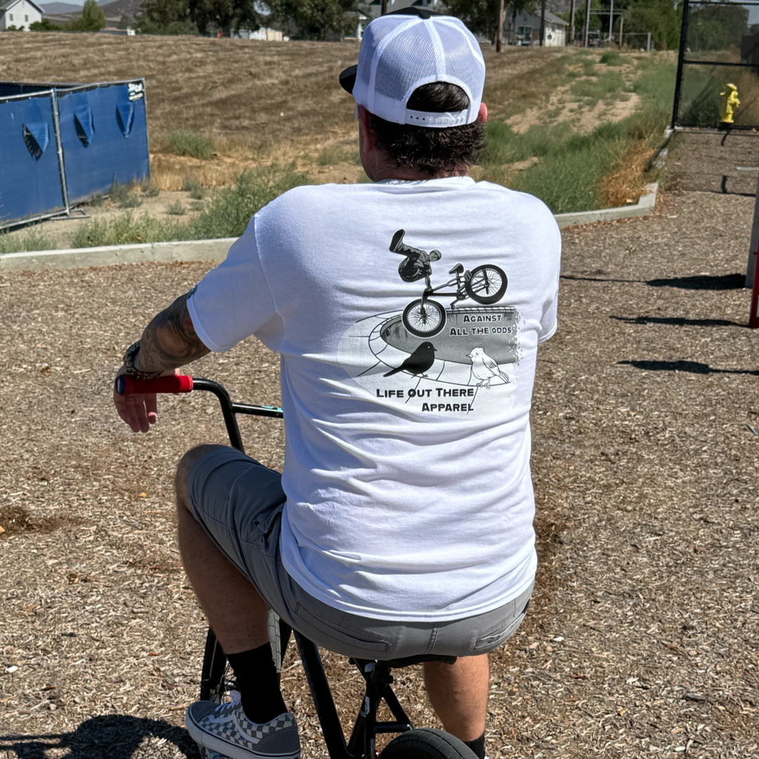 Channeling BMX legend John Buultjens, a man sits outdoors on a small BMX, wearing the Unisex - Against All The Odds / The Ride Tee in white by Life Out There Apparel, featuring a rider graphic and bold text across the back.