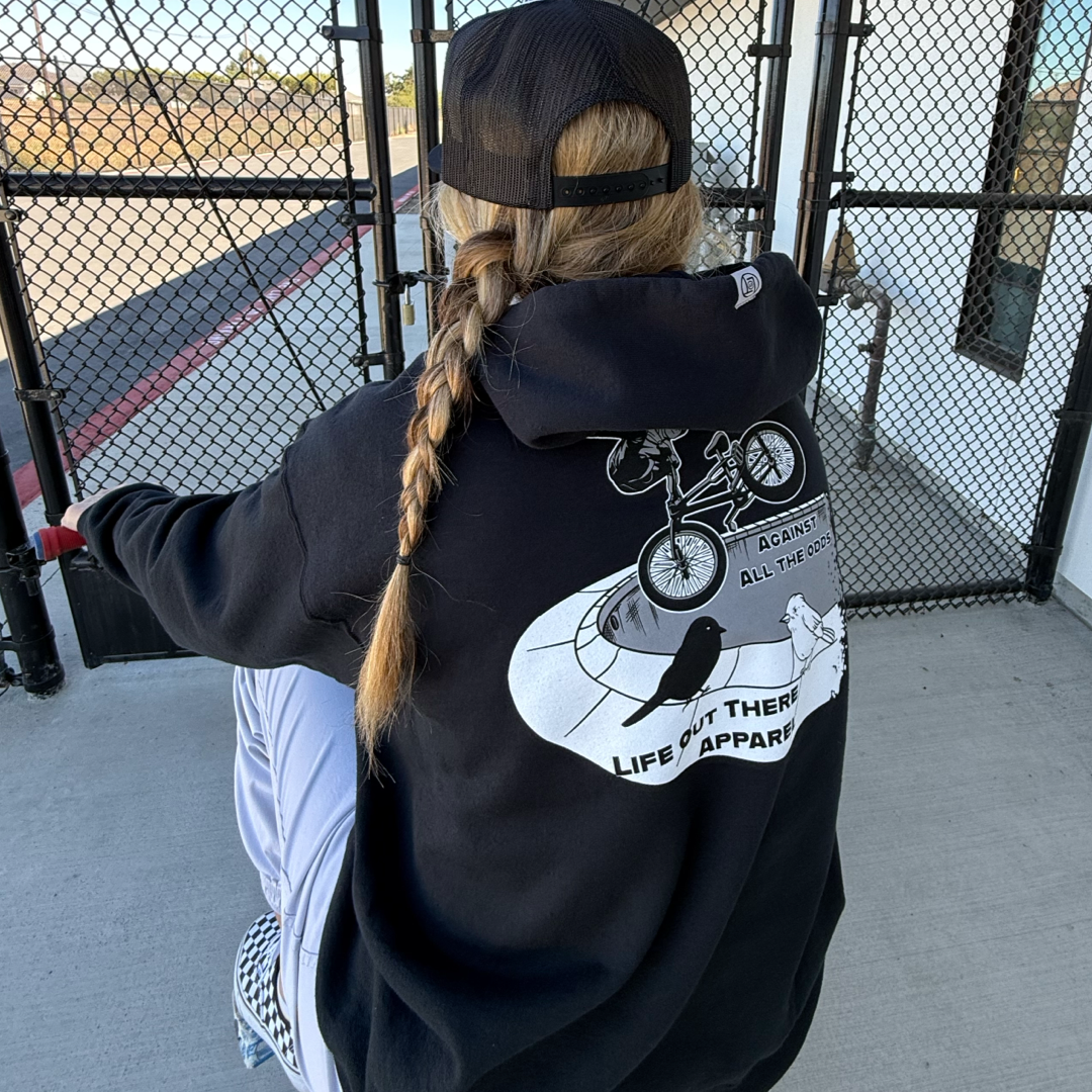 A BMX rider with a long braid, black cap, and checkered shoes sits by a chain-link fence wearing the Life Out There Apparel Unisex Against All The Odds/The Ride Hoodie in black—truly capturing the AGAINST ALL THE ODDS vibe.