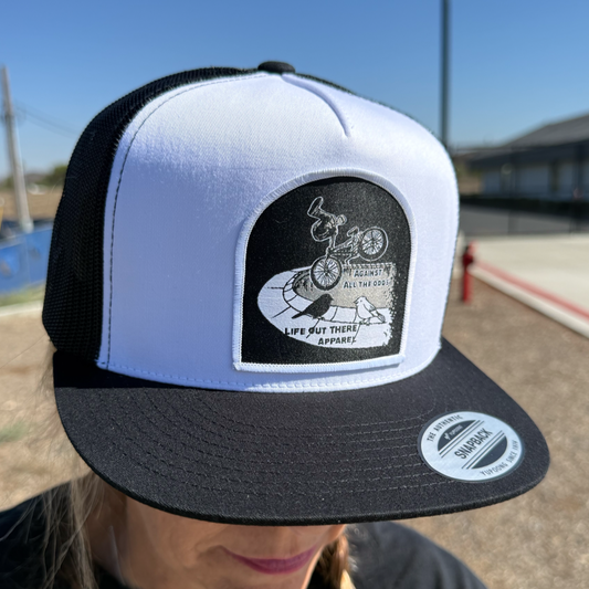 A person wears the “Against All The Odds / The Ride” flat bill snapback trucker hat in white/black by Life Out There Apparel, inspired by John Buultjens, featuring a BMX cyclist patch and a brim sticker.