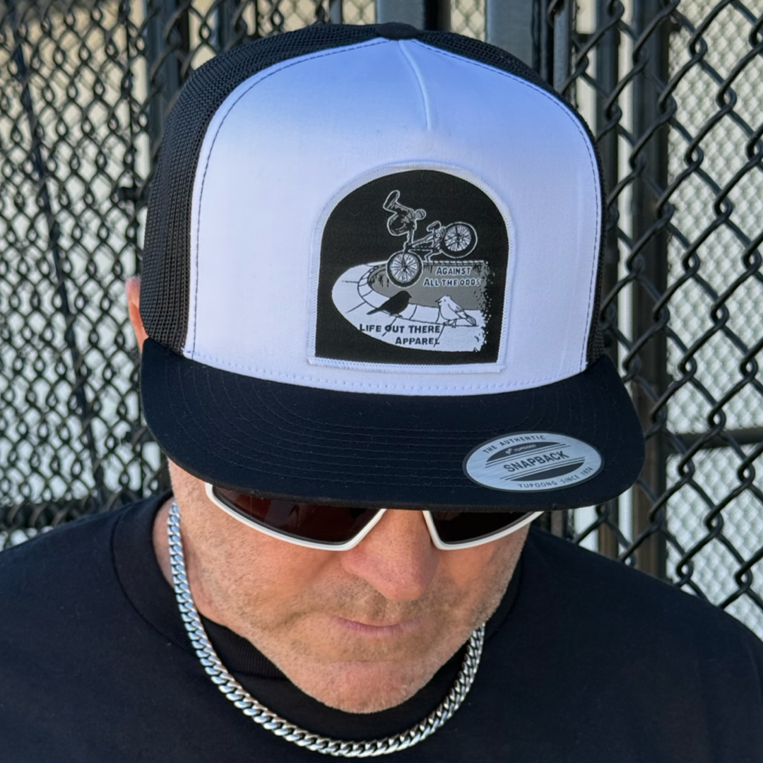 Man wearing sunglasses, a silver chain, and the Against All The Odds / The Ride Flat Bill SnapBack Trucker Hat in white/black by Life Out There Apparel, featuring a BMX patch, stands in front of a metal fence.