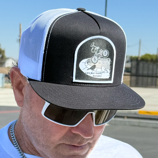 A man wears the Life Out There Apparel Against All The Odds / The Ride flat bill snapback trucker hat in black and white; a chain-link fence and blue sky set a BMX-inspired scene.
