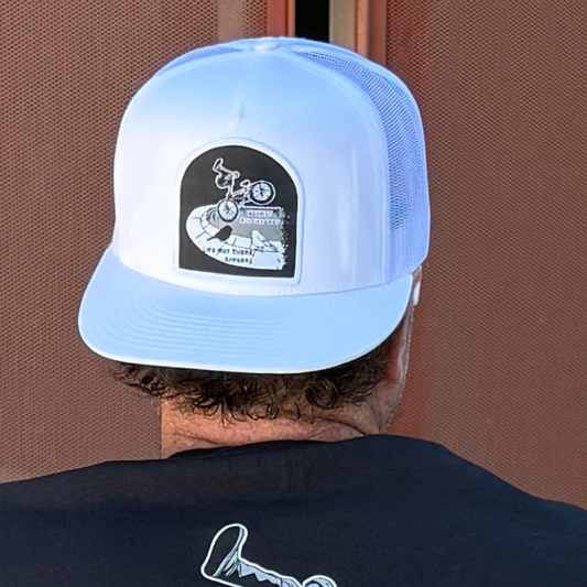 A person seen from behind wears the Life Out There Apparel “Against All The Odds / The Ride” flat bill white SnapBack trucker hat, featuring a black and white graphic of BMX rider John Buultjens above “Cherry Creek Off Road” and “Est. 2021.”.