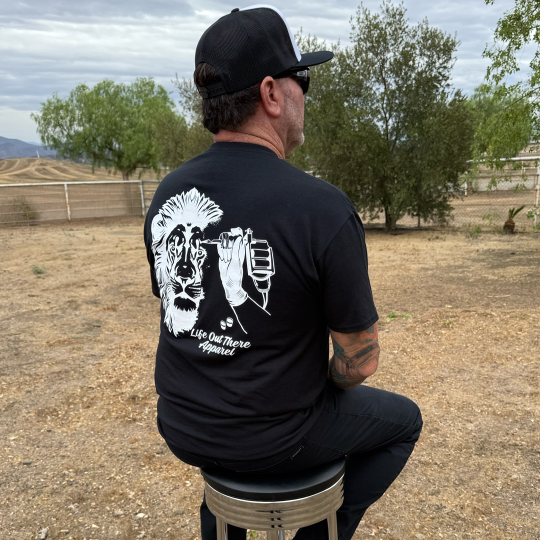 A man wearing a black cap and the Life Out There Apparel Unisex - Not All There Tee in black sits on a stool outdoors near trees and a fenced dirt field.