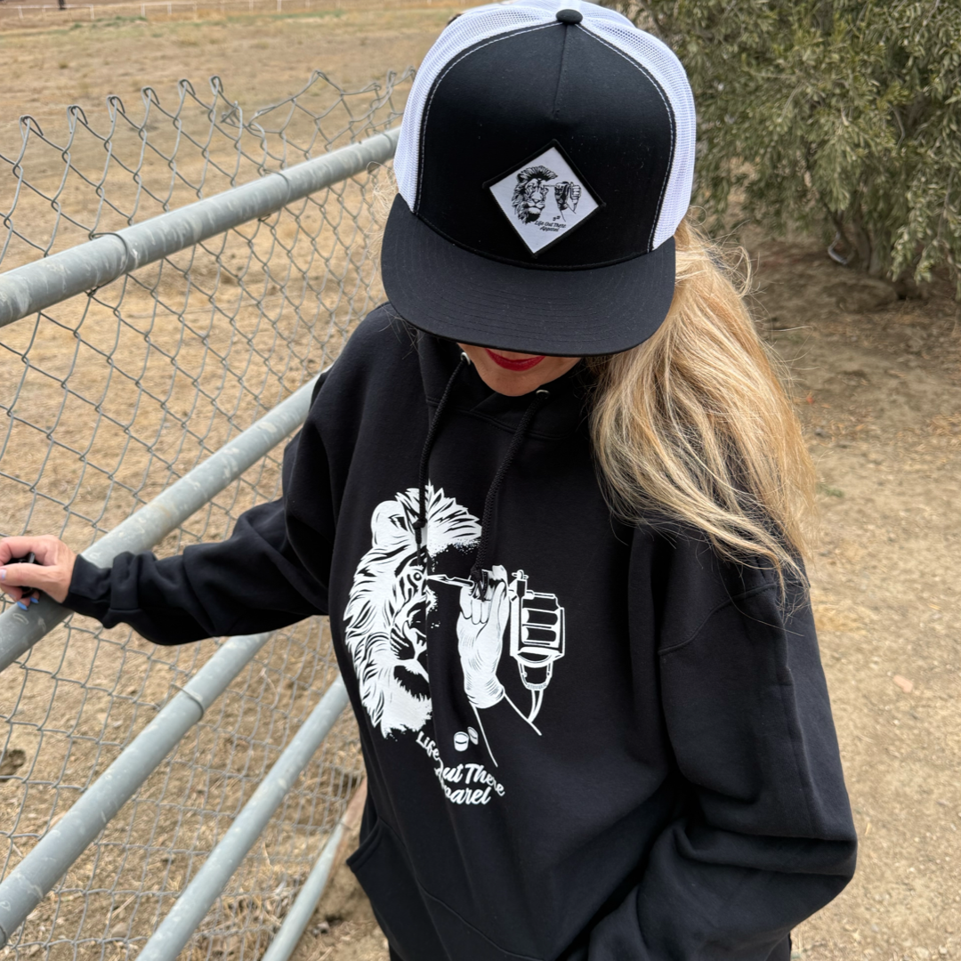 A person with long blonde hair wears the Life Out There Apparel Unisex - Not All There Hoodie in black, made of soft cotton and polyester, styled outdoors by a chain-link fence, paired with a black-and-white cap.