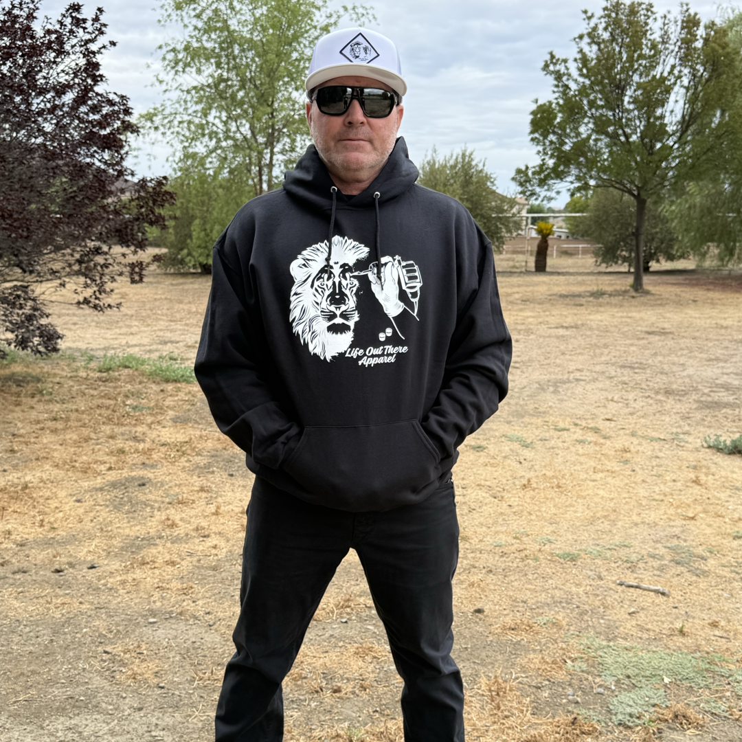 A man wears the Life Out There Apparel Unisex - Not All There Hoodie in black, paired with black pants, sunglasses, and a white cap while standing outdoors on dry grass with trees and a cloudy sky in the background.