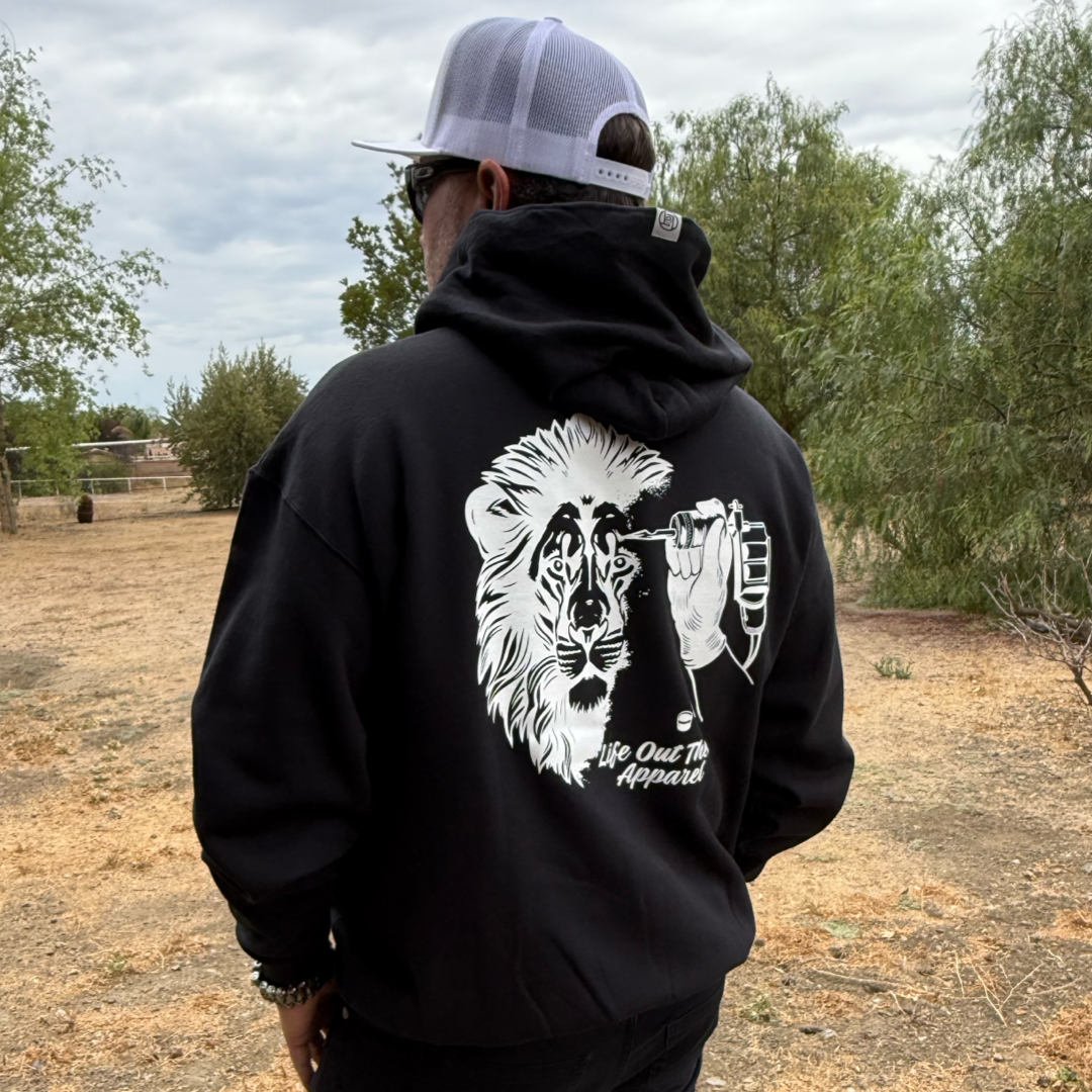 A person wearing the Unisex - Not All There Hoodie in black by Life Out There Apparel, featuring a white outlined lion’s face with a tattoo machine, stands outdoors among trees and dry grass on a cloudy day.